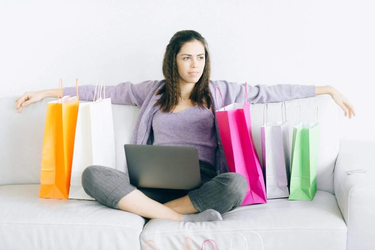 Young woman sitting on a couch with colorful shopping bags and a laptop, representing online shopping.