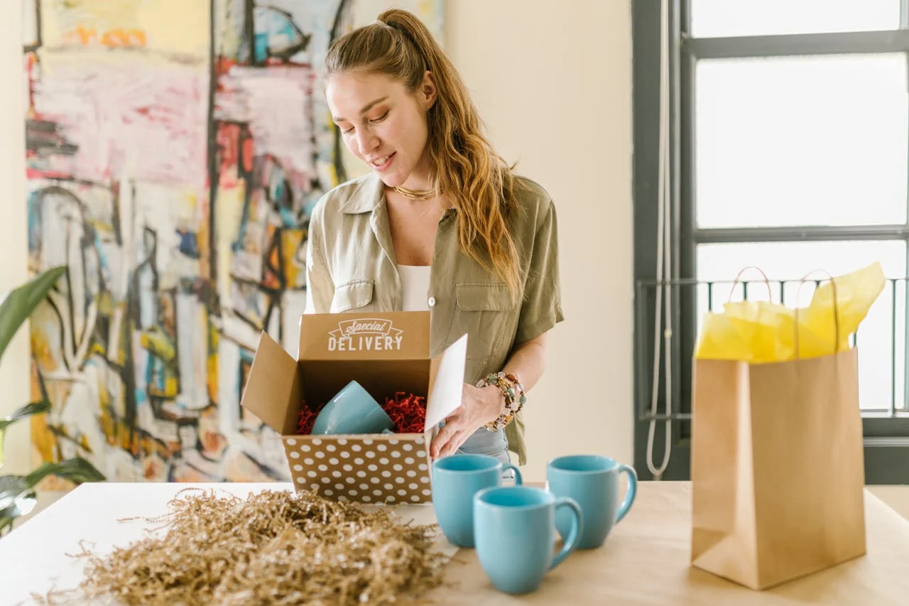Smiling woman unpacking a special delivery box containing blue mugs in a cozy indoor setting.